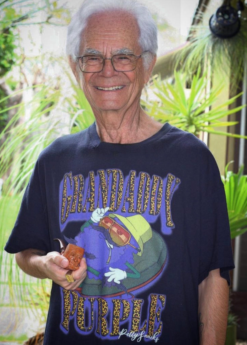 Picture of model wearing Grandaddy Purple t-shirt. The model is and old man smiling, holding a small wooden pipe.