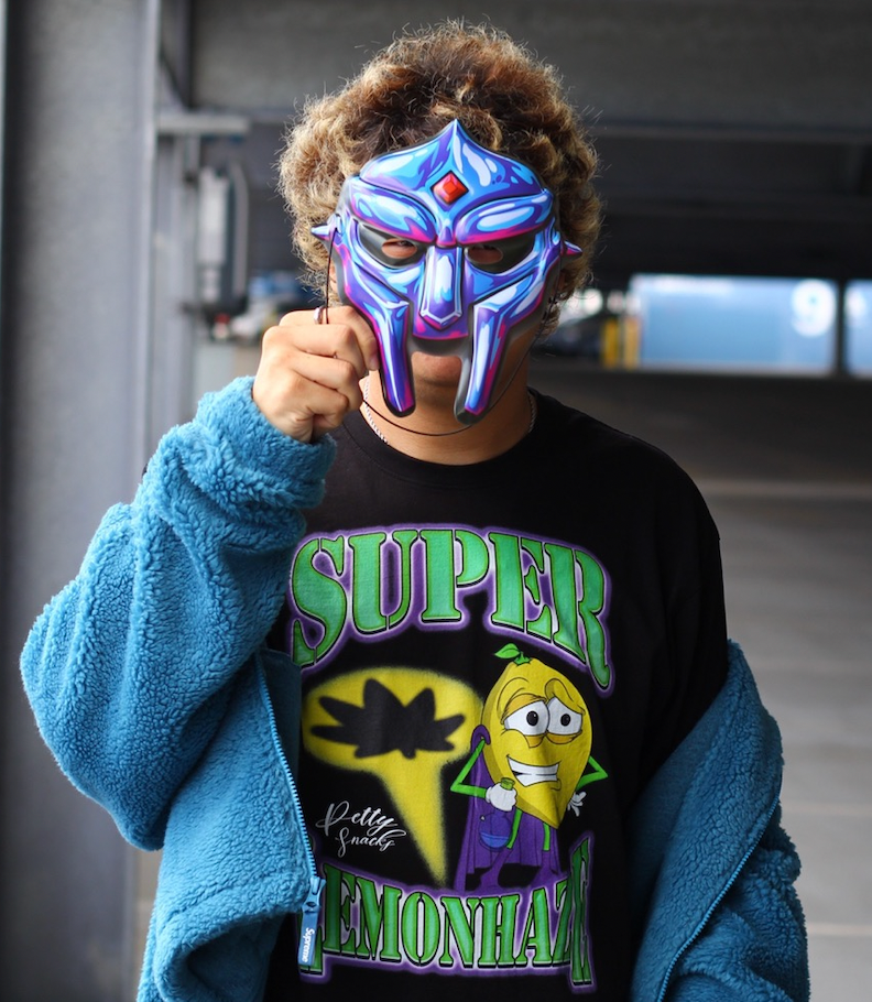 Model pictured. Model wearing black Super Lemon Haze shirt, paired with a aqua green jacket, holding an MF DOOM mask in front of face.